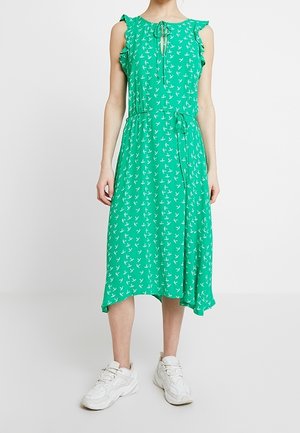 Femme portant une robe midi verte sans manches avec un imprimé d'oiseaux blancs, des épaules à volants, un col à nouer, une ceinture à la taille et des baskets blanches.