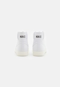 Białe sneakersy na wysokiej cholewce wykonane z materiału płóciennego, z płaską białą podeszwą i dwoma czarnymi logo "NIKE" z tyłu.