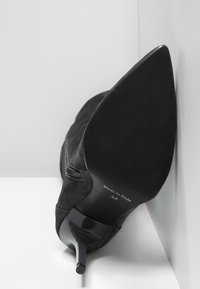 Zapato de tacón alto de ante negro con punta afilada, tacón negro brillante y estampado visible de talla y origen en la suela.