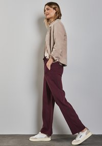 Veste en daim beige, haut gris clair, pantalon large bordeaux et baskets blanches avec des accents gris. Le modèle pose devant un mur gris.