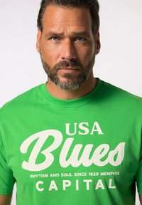 Zelené bavlněné tričko s velkým bílým grafickým potiskem s nápisem "USA Blues" se texturovanými detaily a menším textem popisujícím Memphiskou a rytmus.