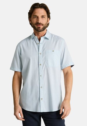 Mann mit dunklem Haar und Bart, der ein hellblaues kurzärmeliges Hemd mit Brusttasche trägt und vor einem einfarbigen Hintergrund steht.
