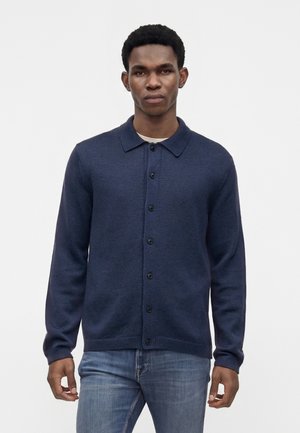 Homme portant un cardigan bleu foncé boutonné par-dessus une chemise blanche et un jean bleu, debout avec les bras détendus devant un fond clair uni.
