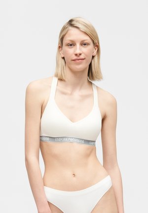 Szőke nő fehér sportmelltartóban, szürke Emporio Armani logós szalaggal és hozzáillő fehér fehérneműben, egyszerű háttér előtt áll.