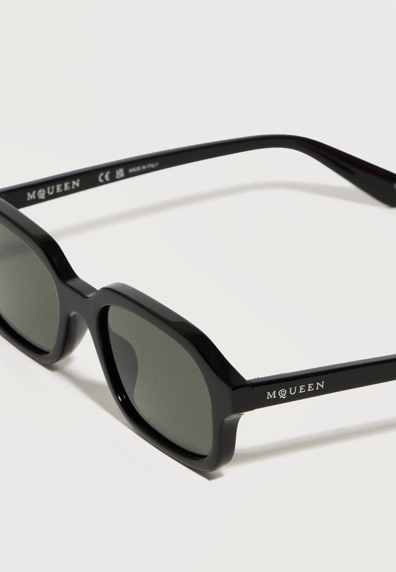 Lunettes de soleil à monture hexagonale noire avec verres foncés et marque "McQueen" sur la branche, posées sur une surface blanche.