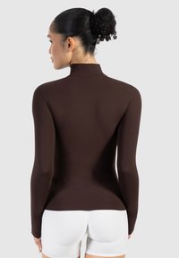 Aansluitend langemouwen top in donkerbruin stof met een hoge kraag. Glad textuur, minimalistisch ontwerp en een op maat gemaakte silhouette.