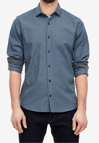 Hombre vestido con una camisa de botones de manga larga azul con patrón y mangas enrolladas, y unos jeans oscuros, de pie con las manos relajadas.