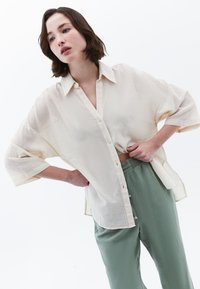 Camicia button-up color crema con tessuto texturizzato e vestibilità ampia, abbinata a pantaloni azzurro chiaro a vestibilità larga.