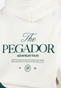 Felpa bianca con cappuccio caratterizzata da scritte verdi, che riportano "The PEGADOR adventure tours" e ulteriori informazioni sul brand in caratteri più piccoli.