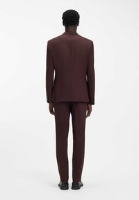 Weinroter Anzug mit maßgeschneiderter Passform, ausgestattet mit einem einzelnen Schlitz auf der Rückseite, schmalen Revers und engen Hosen. Schwarze Schuhe ergänzen den Look.