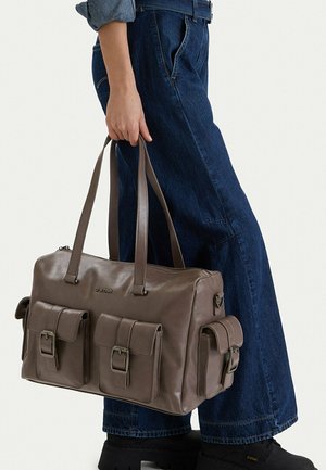 Osoba v modrých džínách držící hnědou koženou kabelku G-Star s třemi kapsami na přezky a dlouhými popruhy.