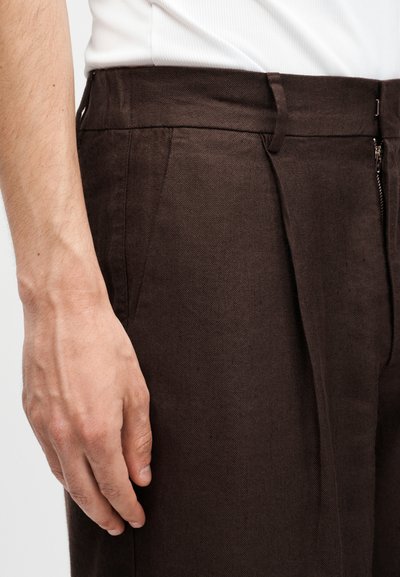 Το αριστερό χέρι του άνδρα ακουμπάει δίπλα σε σκούρο καφέ υφασμάτινο παντελόνι με πιέτες, θηλιές για ζώνη και μερικώς ορατό λευκό πουκάμισο.