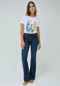 T-shirt blanc avec un motif de Lisbonne, associé à un jean flare bleu foncé. Le modèle se tient debout, mettant en valeur le tissu doux et la coupe décontractée. Des sandales marron complètent le look.