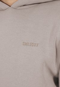 Hooded sweatshirt in lichtbeige stof met een textuurafwerking. Bevat een geborduurd "SMILODOX" logo in een bijpassende tint.