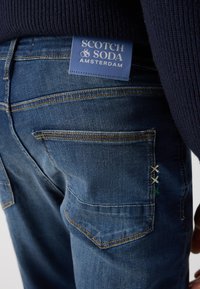 Mørkeblå denim jeans med en blå mærkeetiket, syede lommer og dekorative krydssting i hvidt og grønt.