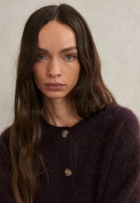 Tmavo fialový, nadýchaný pletený cardigan s okrúhlym výstrihom a tromi veľkými, svetlo hnedými gombíkmi vpredu. Texturovaný, mäkký materiál.