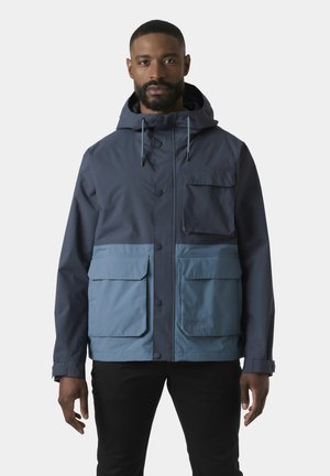 Mężczyzna w ciemnoniebieskiej i jasnoniebieskiej kurtce z kapturem z trzema dużymi przednimi kieszeniami, stojący na tle jednolitego białego tła.