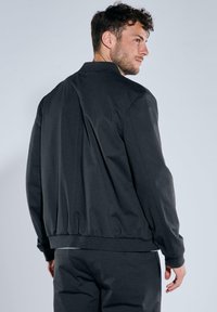 Schwarze Bomberjacke mit glatter Textur, elastischem Bund und minimalistischen Design. Sie verfügt über einen schlichten Kragen und keine sichtbaren Verschlüsse.