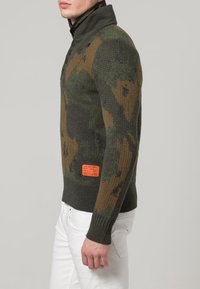 Gestrickter Pullover in grün-braunem Tarnmuster, mit hohem Kragen und strukturiertem Stoff. Orangenes Etikett mit Markenlogo an der unteren Seite.