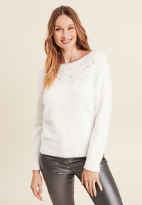 Weißer, flauschiger Pullover mit weitem Ausschnitt, der funkelnde Perlendetails über der Brust aufweist. Kombiniert mit metallisch grauen, eng anliegenden Hosen.
