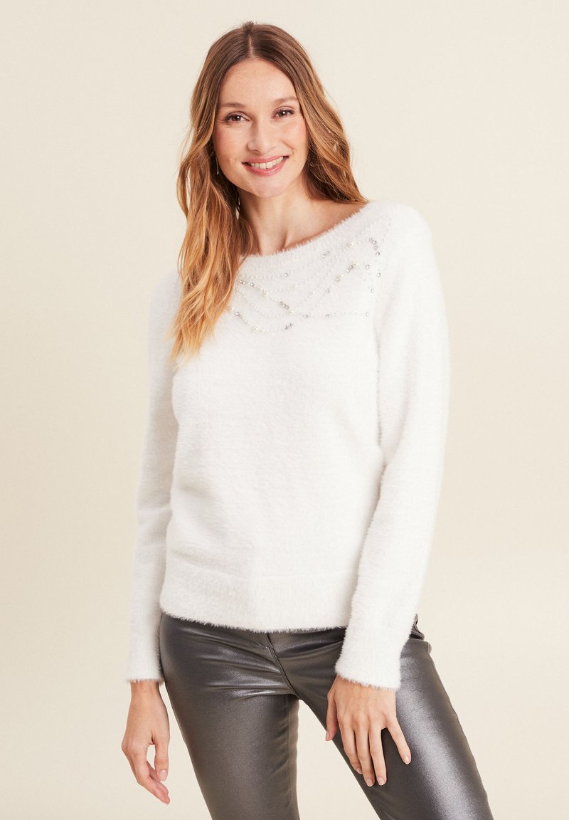 Weißer, flauschiger Pullover mit weitem Ausschnitt, der funkelnde Perlendetails über der Brust aufweist. Kombiniert mit metallisch grauen, eng anliegenden Hosen.