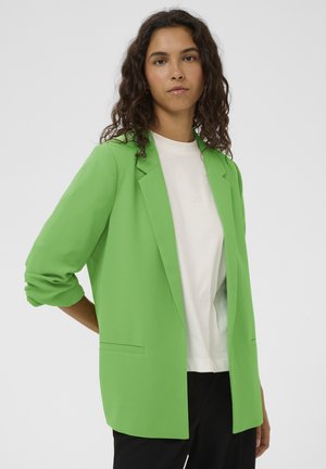 Zeleni blazer iz lahkega materiala, s kljunastim ovratnikom, dvema sprednjima žepoma in zavihanimi rokavi. Nošen čez belo srajco.