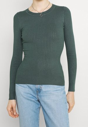 Woman wearing a fitted dark green ribbed long-sleeve sweater and light blue jeans with a silver heart pendant necklace.