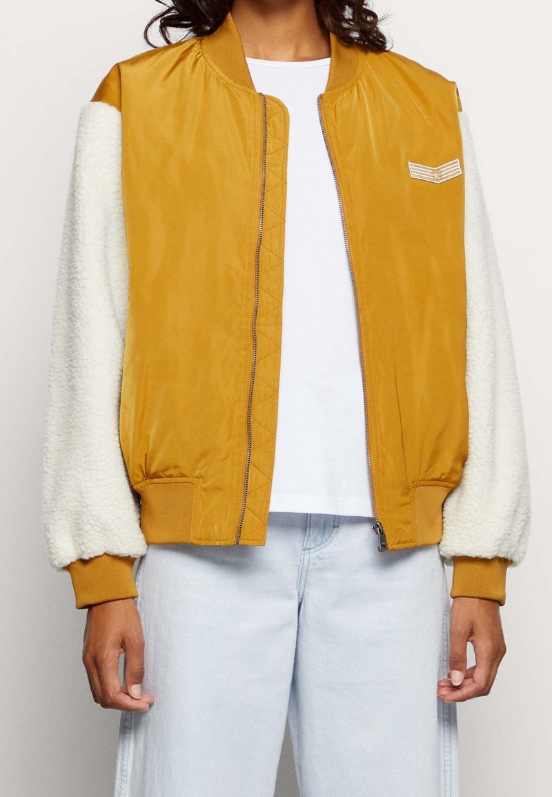 Personne portant une veste bomber jaune moutarde avec des manches blanches duveteuses, une chemise blanche et un jean bleu clair, debout les bras détendus.