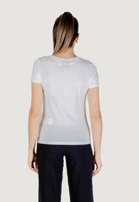 Camiseta blanca de manga corta con cuello redondo, hecha de tela suave. Presenta un sutil logo de EA7 en la parte superior de la espalda. Combinada con pantalones oscuros.