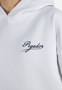 Hanorac alb din amestec de bumbac, cu un logo brodat navy "Pigador" pe piept. Textură netedă, grosime medie.