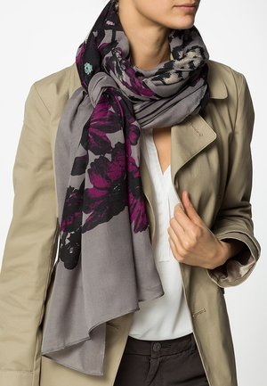 Femme portant un trench-coat beige, un chemisier blanc et une grande écharpe grise avec un imprimé floral violet et noir, lâchement enroulée autour de son cou.
