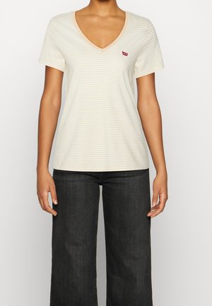 Kobieta ubrana w beżowo-biały pasiasty t-shirt z dekoltem w serek i krótkim rękawem oraz ciemne szerokie jeansy, stoi z rozluźnionymi rękami po bokach.