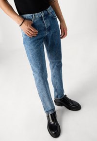 Light blue denim jeans with a straight cut, featuring a classic five-pocket design and subtle fading. Worn with black leather shoes.