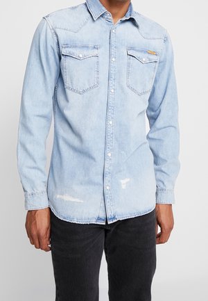 Mężczyzna noszący jasnoniebieską koszulę jeansową z dwoma kieszeniami na piersi i subtelnymi przetarciami, w połączeniu z czarnymi spodniami.