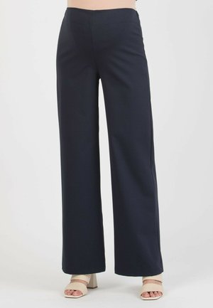 Kobieta w czarnych szerokich spodniach i beżowych sandałach na klocku z odkrytymi palcami, stojąca na tle jednolitego jasnego tła.