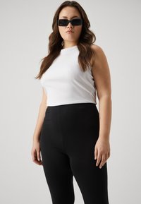 White sleeveless top with a ribbed texture paired with high-waisted black leggings. Model wears black rectangular sunglasses.
