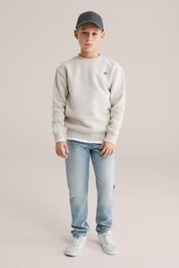 Světlounce šedá texturovaná mikina se šestiúhelníkovým vzorem, v kombinaci se světle modrými džínami a bílými teniskami. Outfit doplňuje šedá čepice.