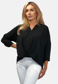 Czarna bluzka z dekoltem w kształcie litery V i luźnym fasonem, z rękawami o długości trzech ćwiartek oraz zawiązanym przodem. Połączona z białymi spodniami.