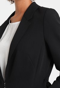 Schwarzer maßgeschneiderter Blazer mit scharfen Revers, strukturierten Schultern und seitlichen Taschen, getragen über einem weißen Oberteil. Verfügt über klare Linien und eine glatte Textur.