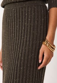 Jupon à côtes vert foncé avec des éclats dorés ; présente une coupe ajustée et une taille élastique. Orné à la main de bracelets en or.