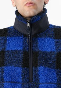 Fleece pullover i blå og sort ternet mønster. Har høj krave, lynlås lukning og tekstureret materiale med en blød finish.