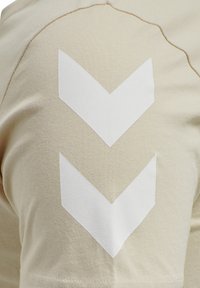 Beige t-shirt lavet af bomuld, med to hvide chevron-mønstre på venstre skulder. Glat tekstur med synlige syningsdetaljer.
