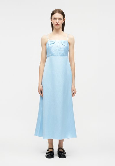 Γυναίκα που φοράει αμάνικο midi φόρεμα ανοιχτού γαλάζιου χρώματος με ρεβέρ στο μπούστο και μαύρα παπούτσια με αγκράφα, στέκεται μπροστά σε ένα απλό φόντο.