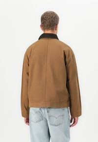 Mann som vender bort med en brun jakke med svart krage og lyseblå jeans mot en ensfarget hvit bakgrunn.