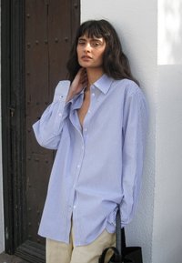 Blue and white striped button-up shirt with oversized sleeves and a deep neckline, paired with light beige pants and a black bag.