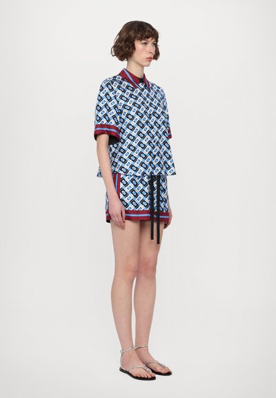 Cămașă scurtă cu mâneci, cu un model albastru și accente roșii, asociată cu pantaloni scurți asortați și sfoară neagră. Modelul poartă sandale argintii cu curea.