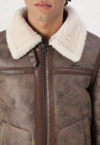 Ruda odinė striukė su kreminio spalvos avikailio apykakle, užtrauktuku, dviem didelėmis priekinėmis kišenėmis ir tekstūruotu paviršiumi.