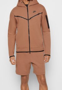 Hnědá mikina Nike se zipem a kapucí, se černými akcenty a boční kapsou. V páru s hnědými kraťasy ve stejném odstínu. Hladká textura.