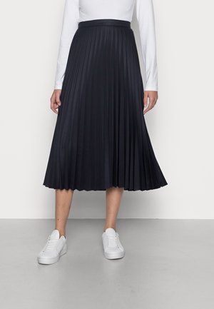 Frau trägt einen schwarzen plissierten Midirock, ein weißes langärmeliges Oberteil und weiße Sneakers, steht auf einem grauen Boden vor einer weißen Wand.