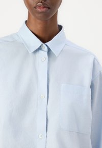 Camicia azzurro chiaro con bottoni, dotata di colletto e tasca sul lato sinistro, realizzata in un tessuto morbido e strutturato. I bottoni sono bianchi.